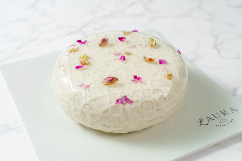 A round cake with white icing, decorated with flower petals, on a plate with the brand name 'LAURA' visible.