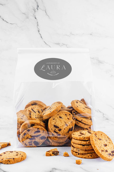 Image of Chocolate Sea-Salt Sablés by Laura's Home Bakery, beautifully presented in their packaging, with rich chocolate chips and delicate sea-salt crystals atop golden-brown cookies.