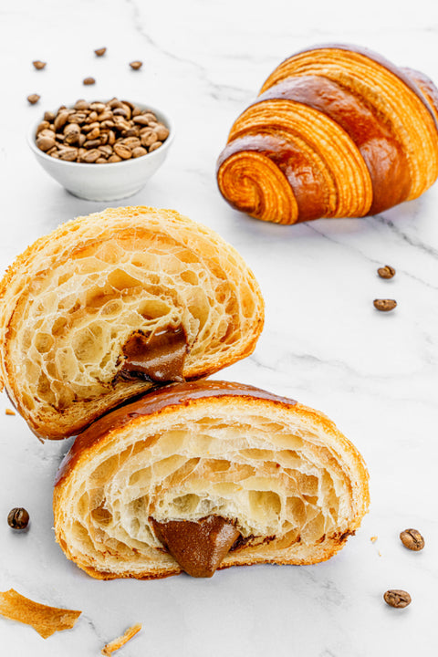 Image of a sliced Coffee Croissant from Laura's Home Bakery, a delectable treat designed to complement the rich flavors of coffee, showcasing its golden-brown, flaky exterior beside coffee beans