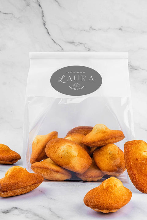 Image of elegantly packaged Madeleines from Laura's Home Bakery in a white bag with a transparent bottom half, featuring the bakery's logo prominently in the center.