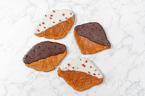 Four flat croissants on a marble surface, two with chocolate topping and two with a cream filling, sprinkled with sugar.