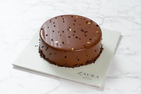 A chocolate cake with a glossy icing and sprinkles on top, presented on a white plate with the brand name 'Laura' inscribed.