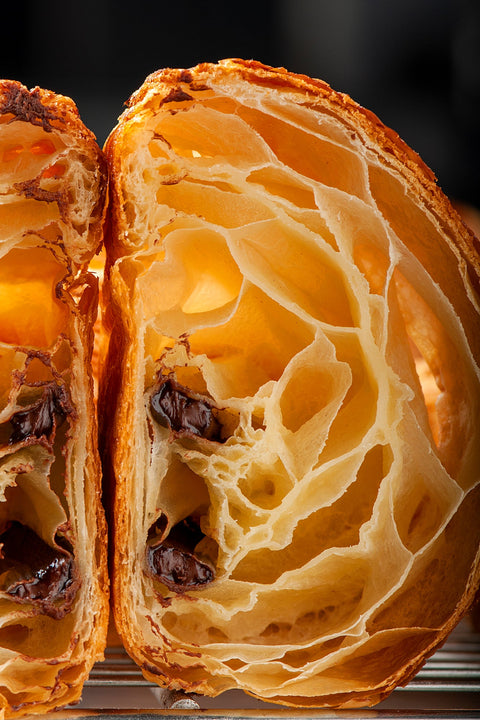Close-up side view of a perfectly baked Pain Au Chocolat from Laura's Home Bakery, displaying its golden-brown, flaky layers and a luscious core of rich, velvety chocolate.