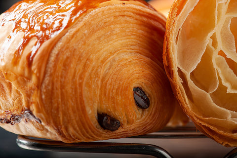 Close-up of a perfectly baked Pain Au Chocolat from Laura's Home Bakery, displaying its golden-brown, flaky layers and a luscious core of rich, velvety chocolate.