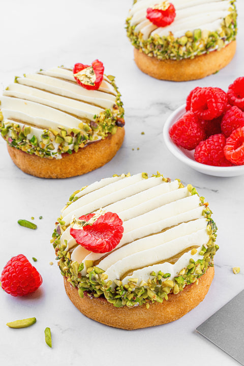 Close-up view of a mini Pistachio and Raspberry Tart from Laura's Home Bakery, highlighting the meticulous arrangement of crushed pistachios and ripe raspberries on a petite, golden pastry crust.